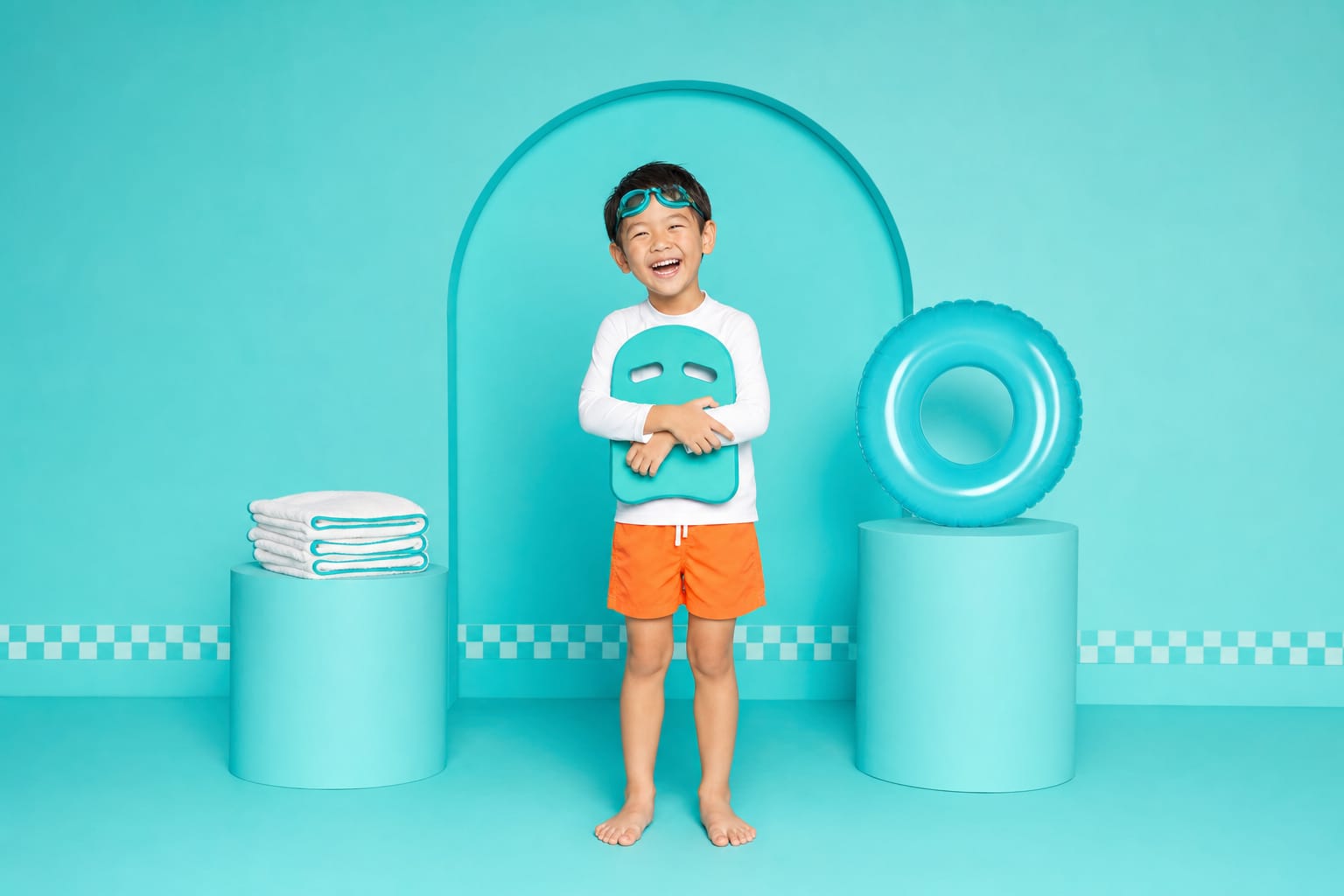 A boy with teal goggles on his forehead hugs a teal kickboard, laughing in an aqua studio.